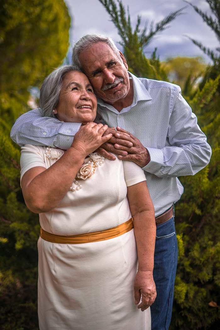 Casal de idosos sorrindo ao ar livre, com o marido abraçando a esposa por trás